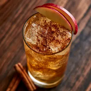 Spiced Apple Rum Cocktail In A Highball Glass With Ice, Apple Fan Garnish, And Cinnamon On Dark Walnut Wood