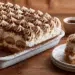 Eggless Tiramisu With Piped Cream Rosettes And Cocoa Dusting In White Dish, Corner Slice On Plate Beside It
