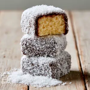 Coconut-Coated Chocolate Lamingtons On A Wire Rack And White Plate, One Halved Showing Golden Sponge Inside